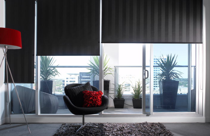 Dark gray roller blinds half open with a red lamp and black chair with red pillows